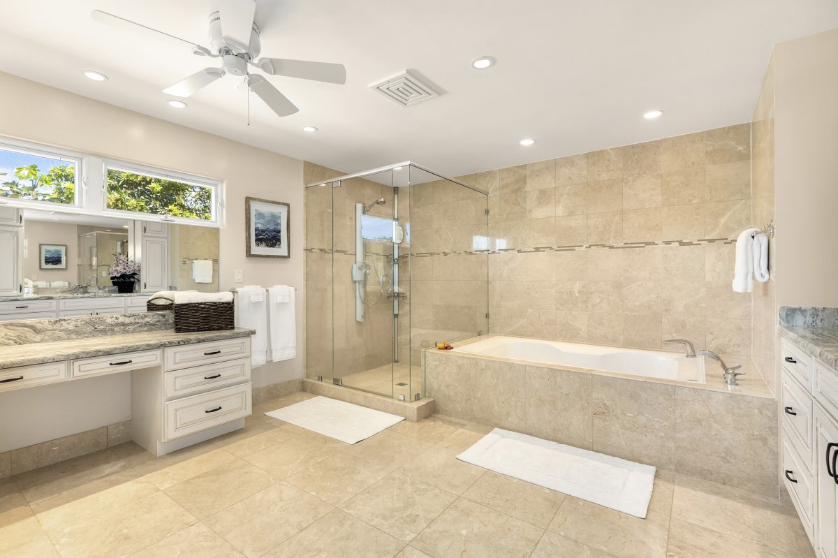 Hale Makana's elegant-looking primary bathroom with a vanity area, ceiling fan, granite countertop, drawers, cabinets, soaking tub and a glass-enclosed walk-in shower