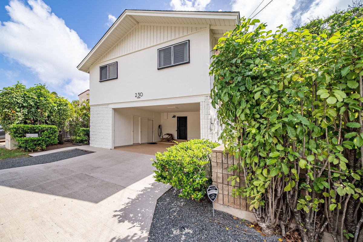 Charming front view of the home featuring a spacious driveway and tropical landscaping, offering a warm island welcome.