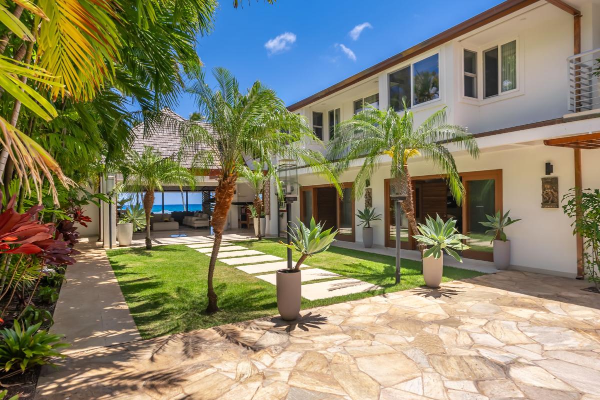 Modern tropical villa framed by lush palm trees and manicured lawns, with a glimpse of turquoise ocean just beyond the property's peaceful grounds.