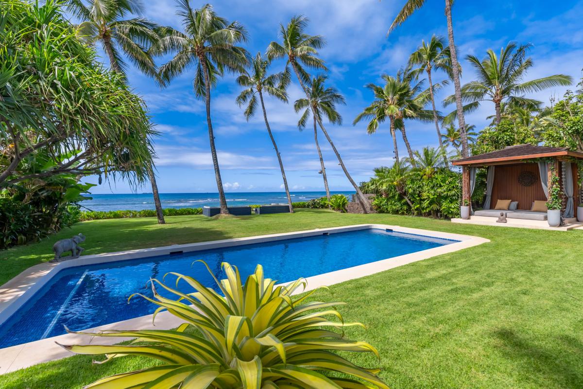 A pristine oceanfront retreat featuring a sparkling pool, lush tropical gardens with swaying palms, and stunning sea views—your perfect Hawaiian escape awaits.