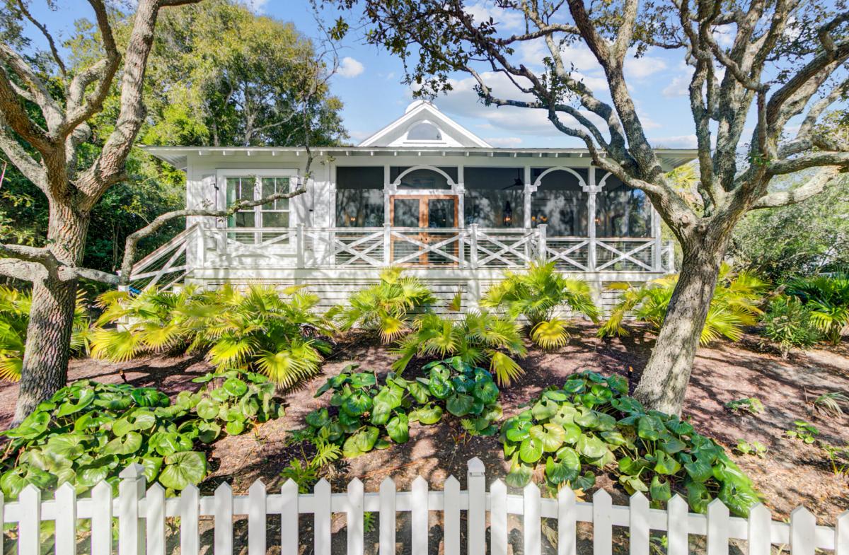 Charming cottage-style property nestled among mature trees with lush tropical landscaping and white picket fence entrance.